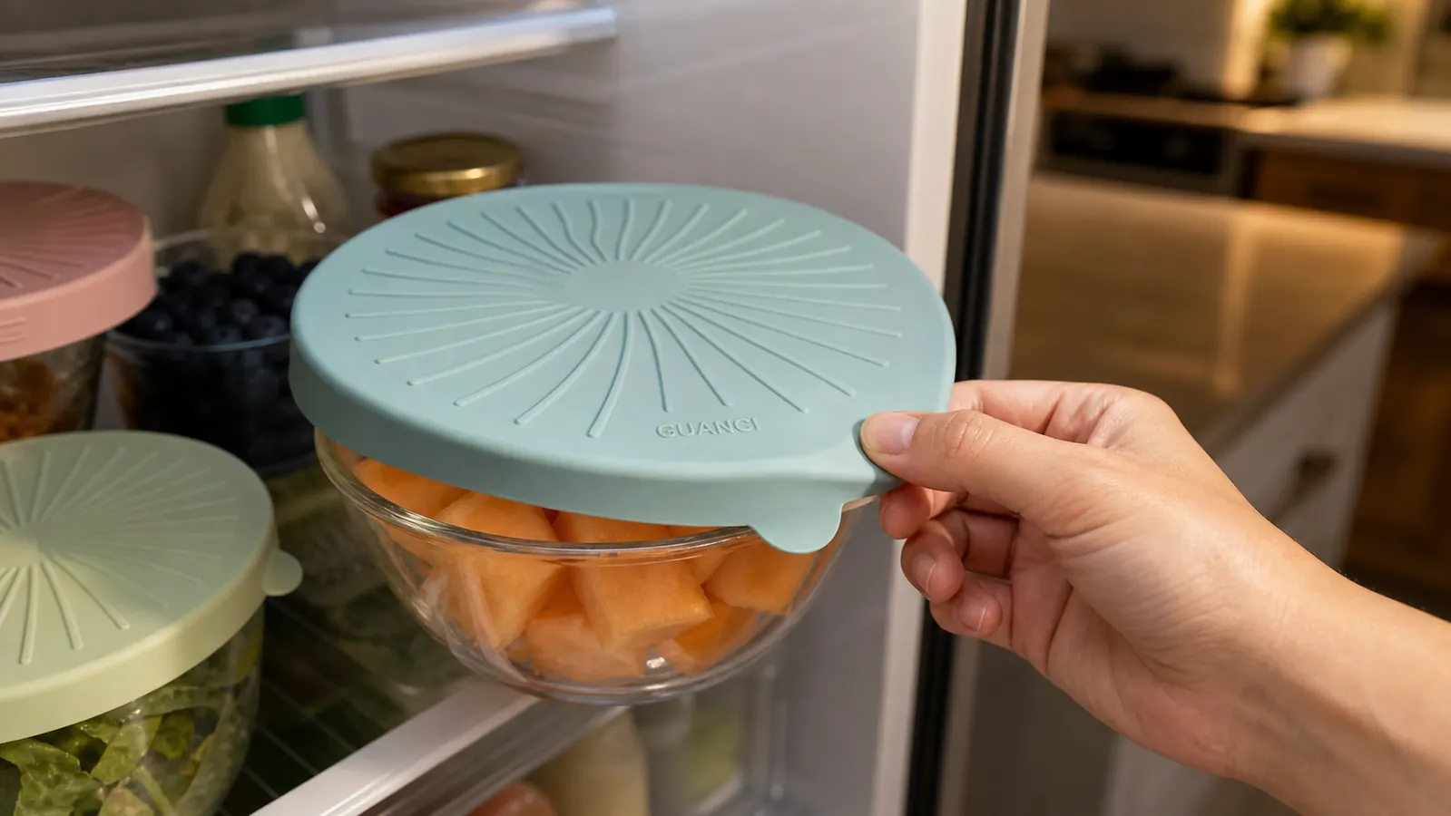 A hand stretching a green silicone lid over a bowl of cut cantaloupe in the refrigerator