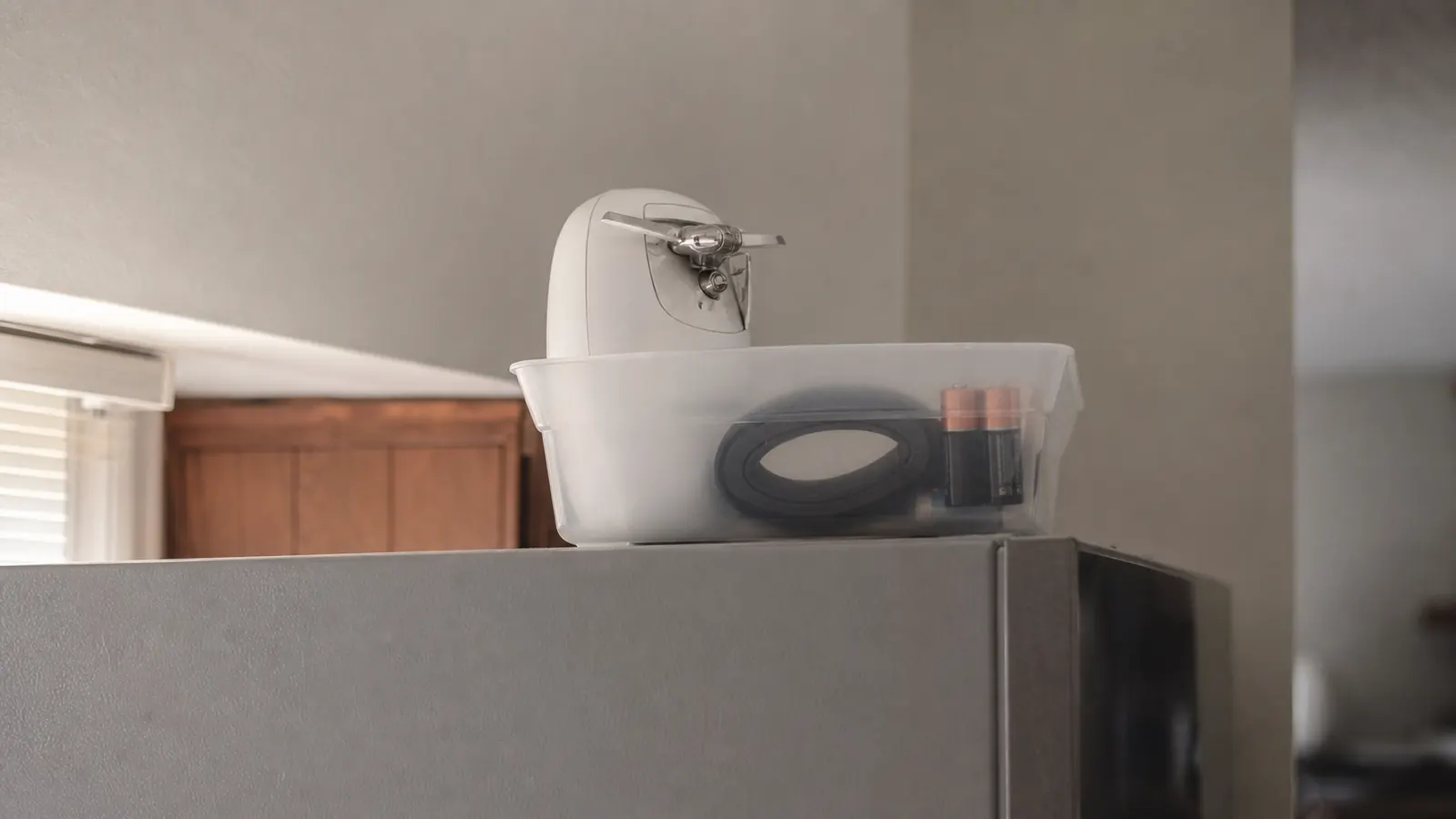 An electric can opener stored in a plastic bin on top of a refrigerator because it did not fit in the kitchen utensil drawer