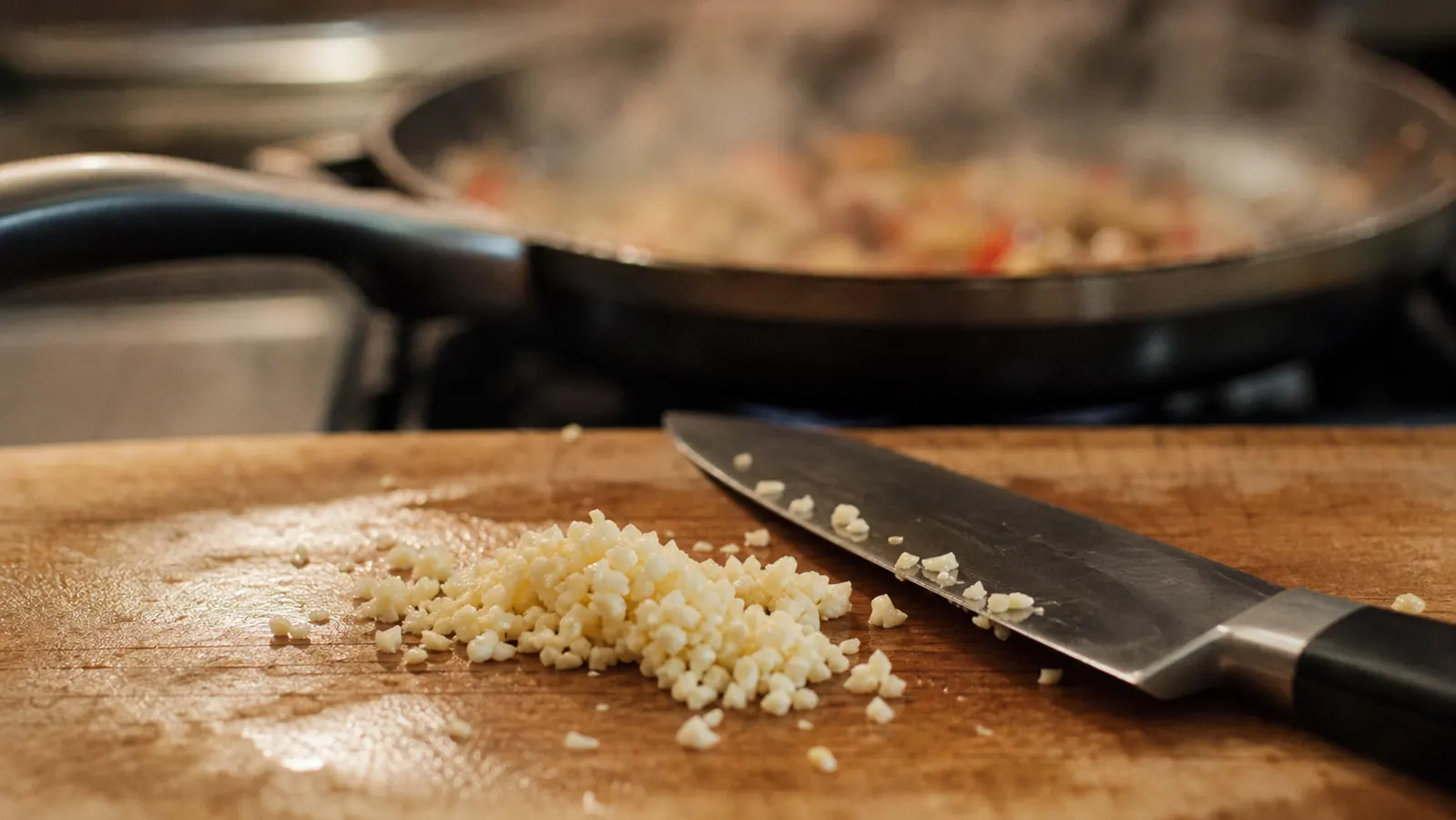 A cutting board with minced garlic and a chef's knife resting beside it, steam rising from a skillet in the background