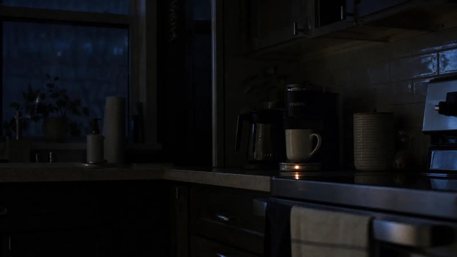 A dark kitchen at night with only the small indicator light on the mug warmer glowing near the coffee maker
