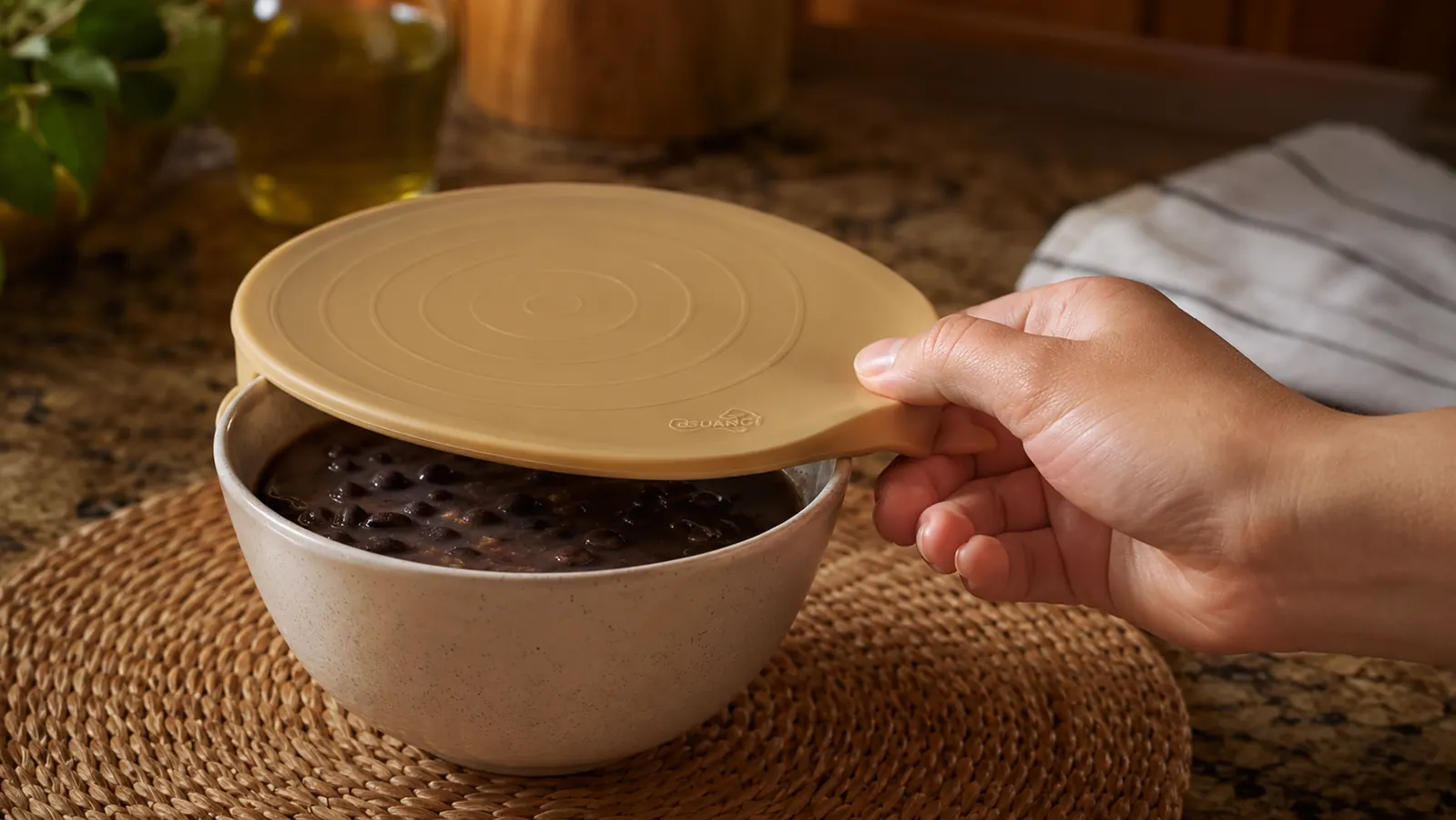A hand stretching a terracotta-colored silicone lid over a bowl of black bean soup on a kitchen counter