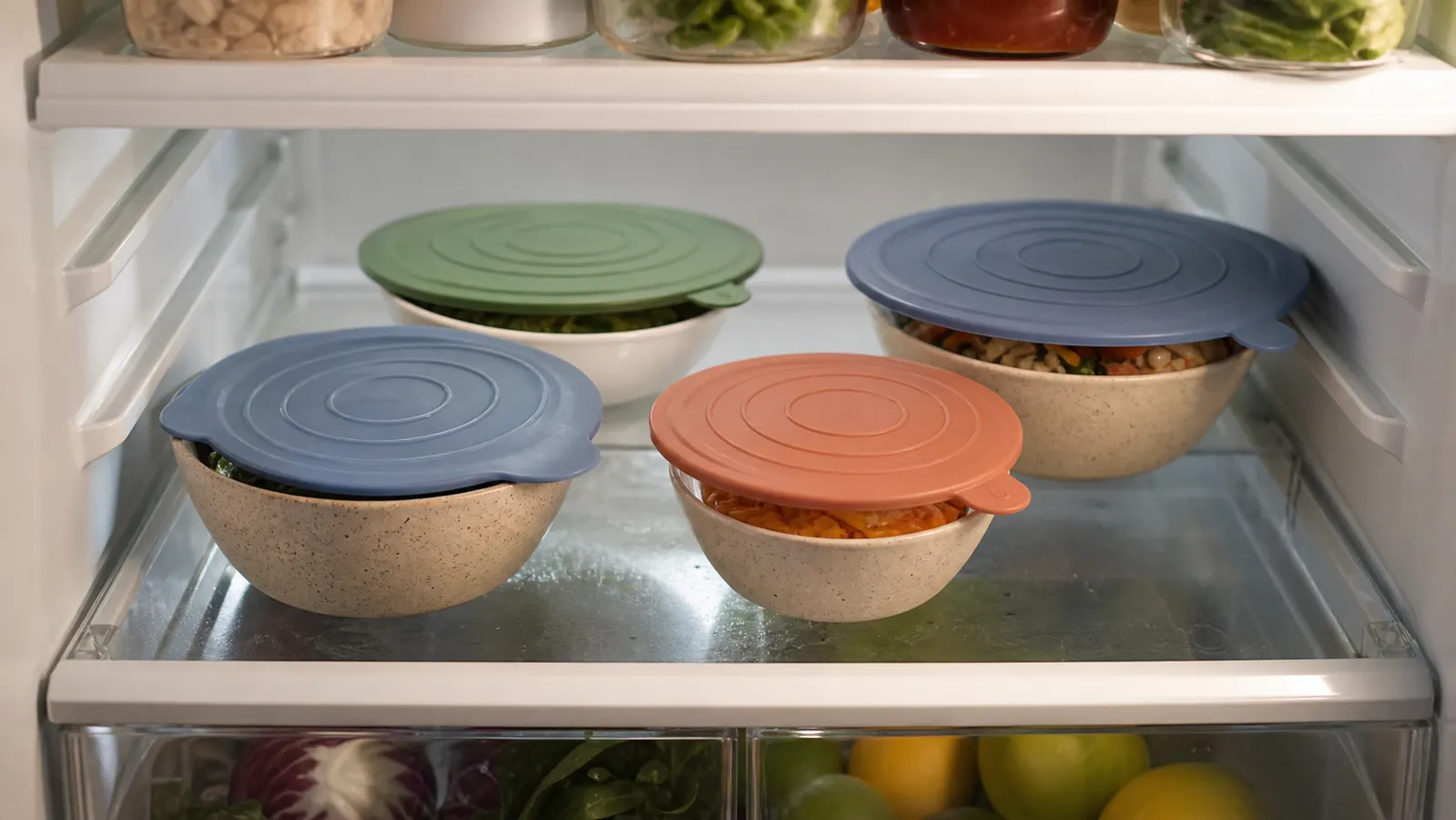 A refrigerator shelf lined with several bowls of covered leftovers, each sealed with a different colored silicone lid