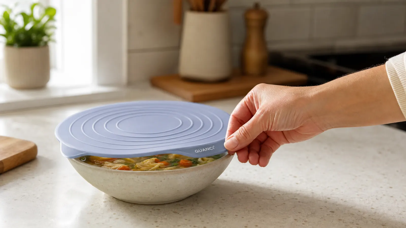 A hand stretching a silicone lid over a round ceramic bowl of leftover soup